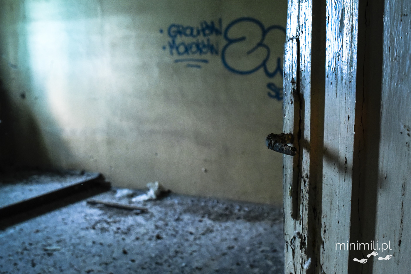 Opuszczony korytarz dawnego szpitala Omega z graffiti i światłem z otwartych drzwi